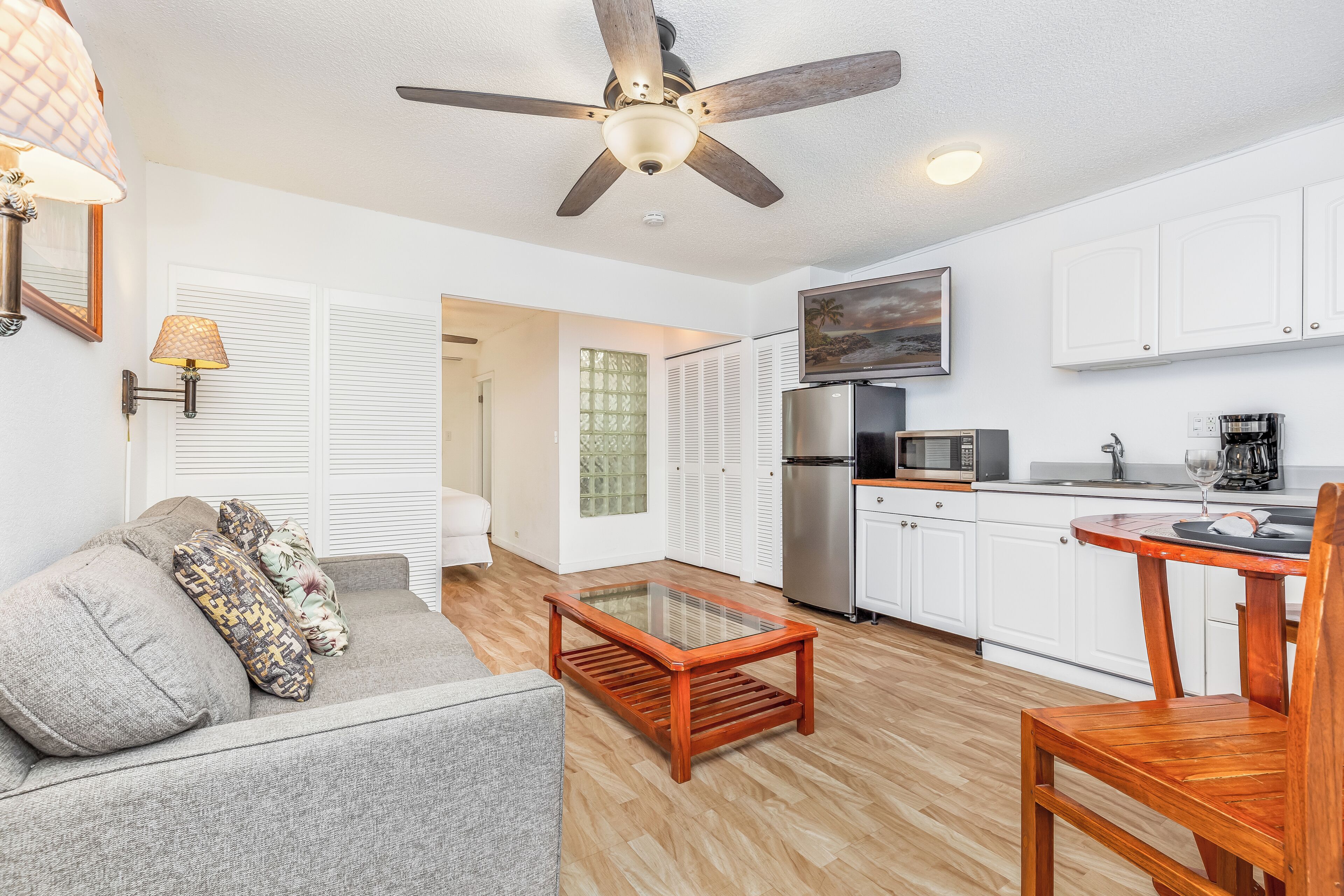 A compact living space with a ceiling fan, a sofa with patterned cushions, a wooden coffee table, a kitchen area with appliances, and a TV mounted above the counter.