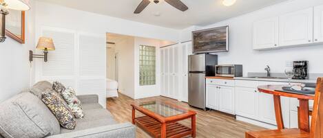 A compact living space with a ceiling fan, a sofa with patterned cushions, a wooden coffee table, a kitchen area with appliances, and a TV mounted above the counter.