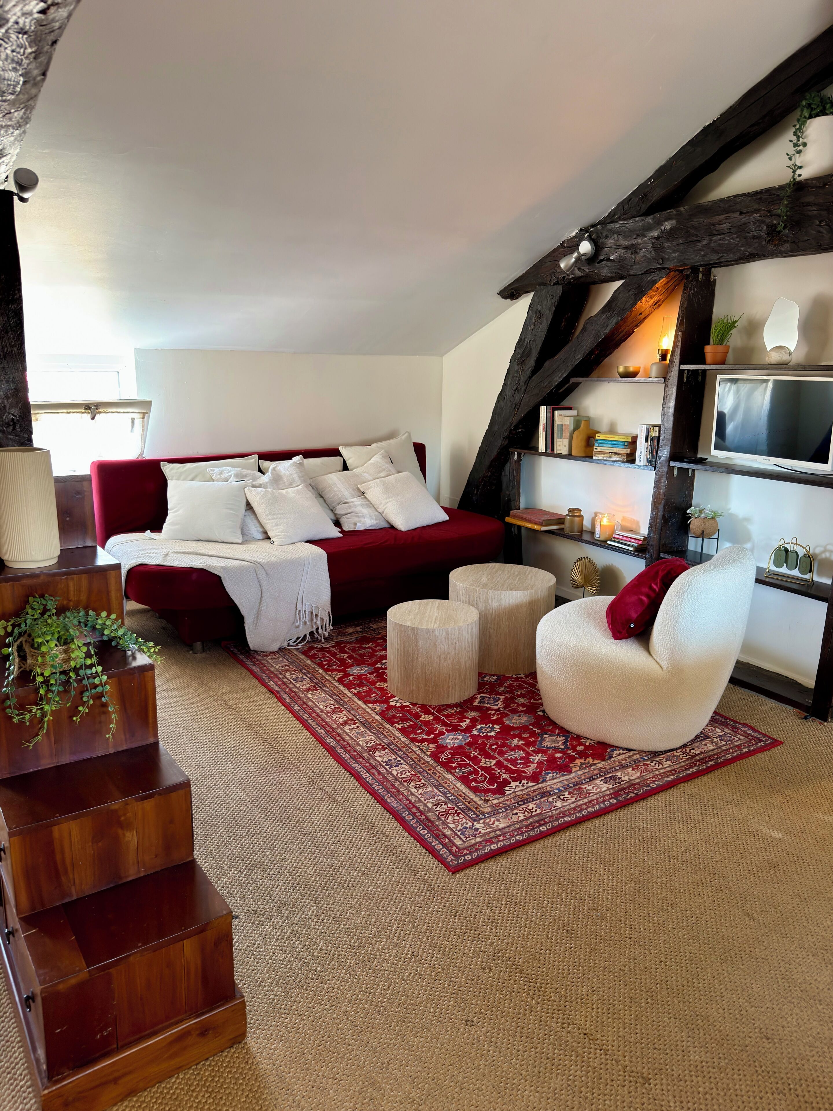A cozy living room with a red sofa, a white armchair, a wooden staircase, and a red patterned rug.