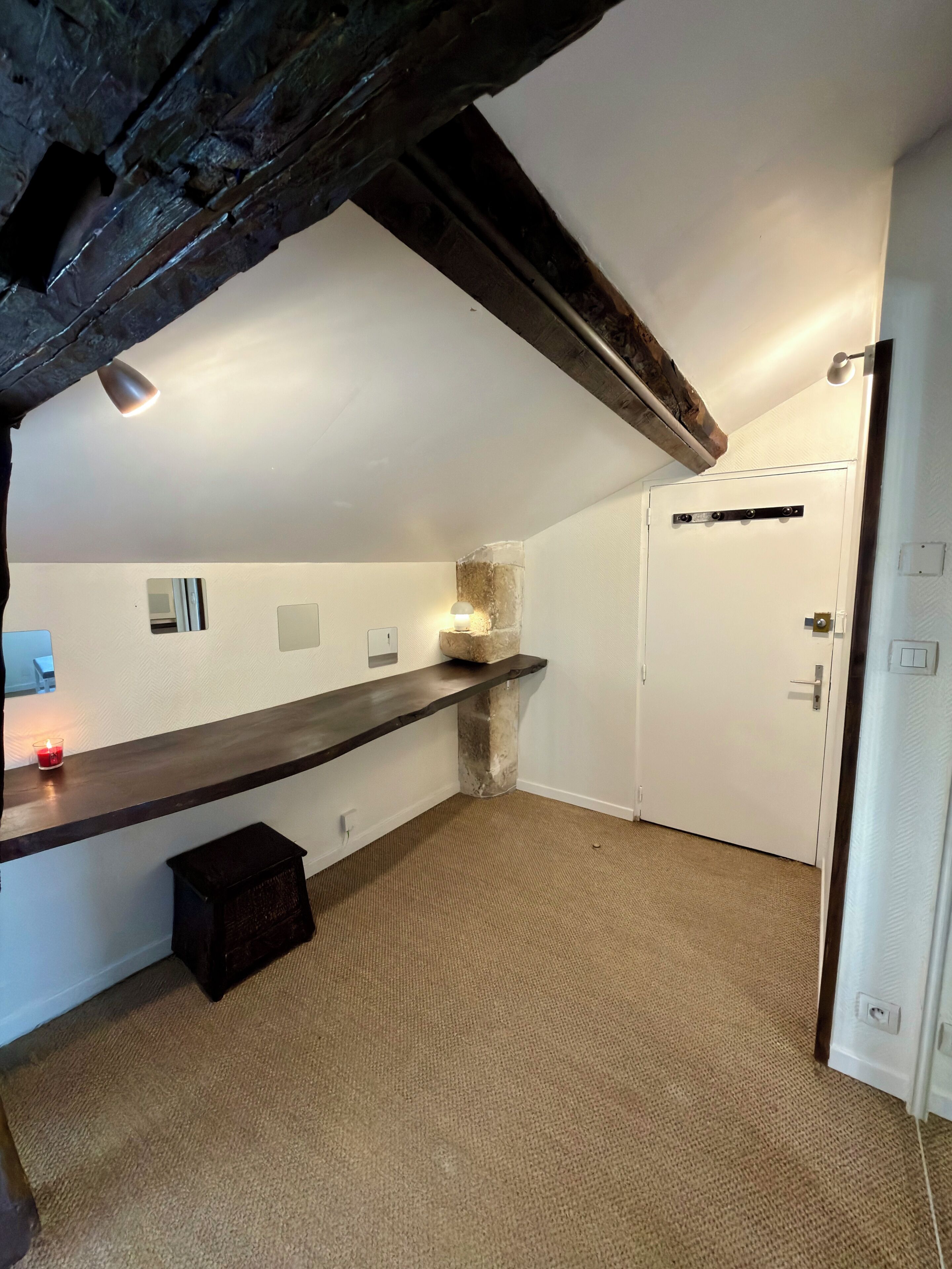 Una habitación con techo inclinado, un banco de madera oscura, una pequeña vela roja, un pilar de piedra y una puerta blanca.