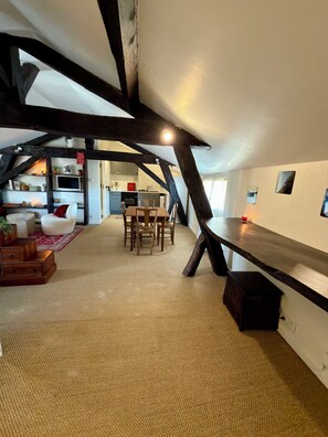 A loft-style room with exposed beams, a dining area with a table and chairs, and a kitchen in the background.