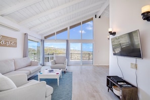 A spacious living room with a high ceiling, large windows, a flat-screen TV, and a comfortable seating area.