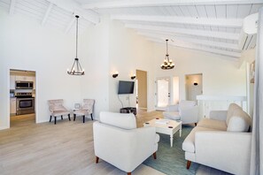 A spacious living room with a kitchen in the background, featuring modern furniture and a high ceiling with exposed beams.