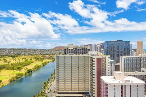 A high-rise building with multiple floors and balconies, situated next to a river with a park and residential area in the background. - Ocean-golf Course Couples Retreat at Island Colony Short Walk to the Beach (Honolulu)