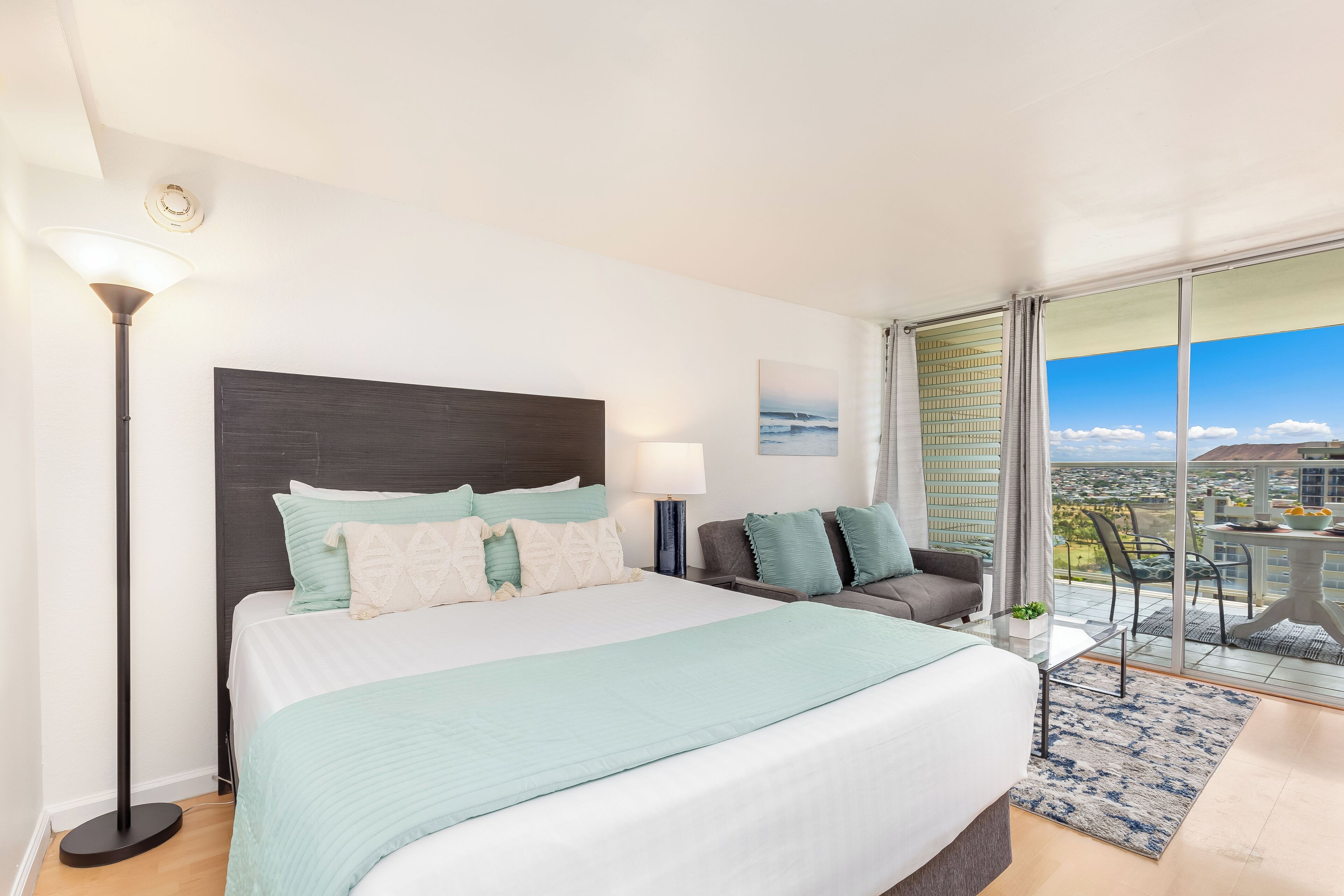 Habitación de hotel con una cama grande, una mesita de noche, una lámpara, vistas a la ciudad y un balcón con mesa y sillas.