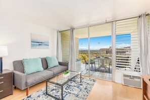 A modern living room with a grey sofa, a glass coffee table, and a view of a cityscape through large sliding glass doors. - Ocean-golf Course Couples Retreat at Island Colony Short Walk to the Beach (Honolulu)