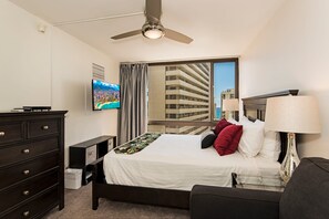 A hotel room with a bed, a dresser, a TV, a window with a view of a building, and a ceiling fan. - Ocean & Diamond Head Views Condo at Waikiki Sunset Short Walk to the Beach (Honolulu)