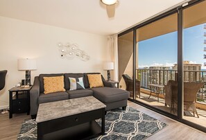A modern hotel room with a grey sectional sofa, a small table, two nightstands, and a large balcony with wicker furniture. - Ocean & Diamond Head Views Condo at Waikiki Sunset Short Walk to the Beach (Honolulu)