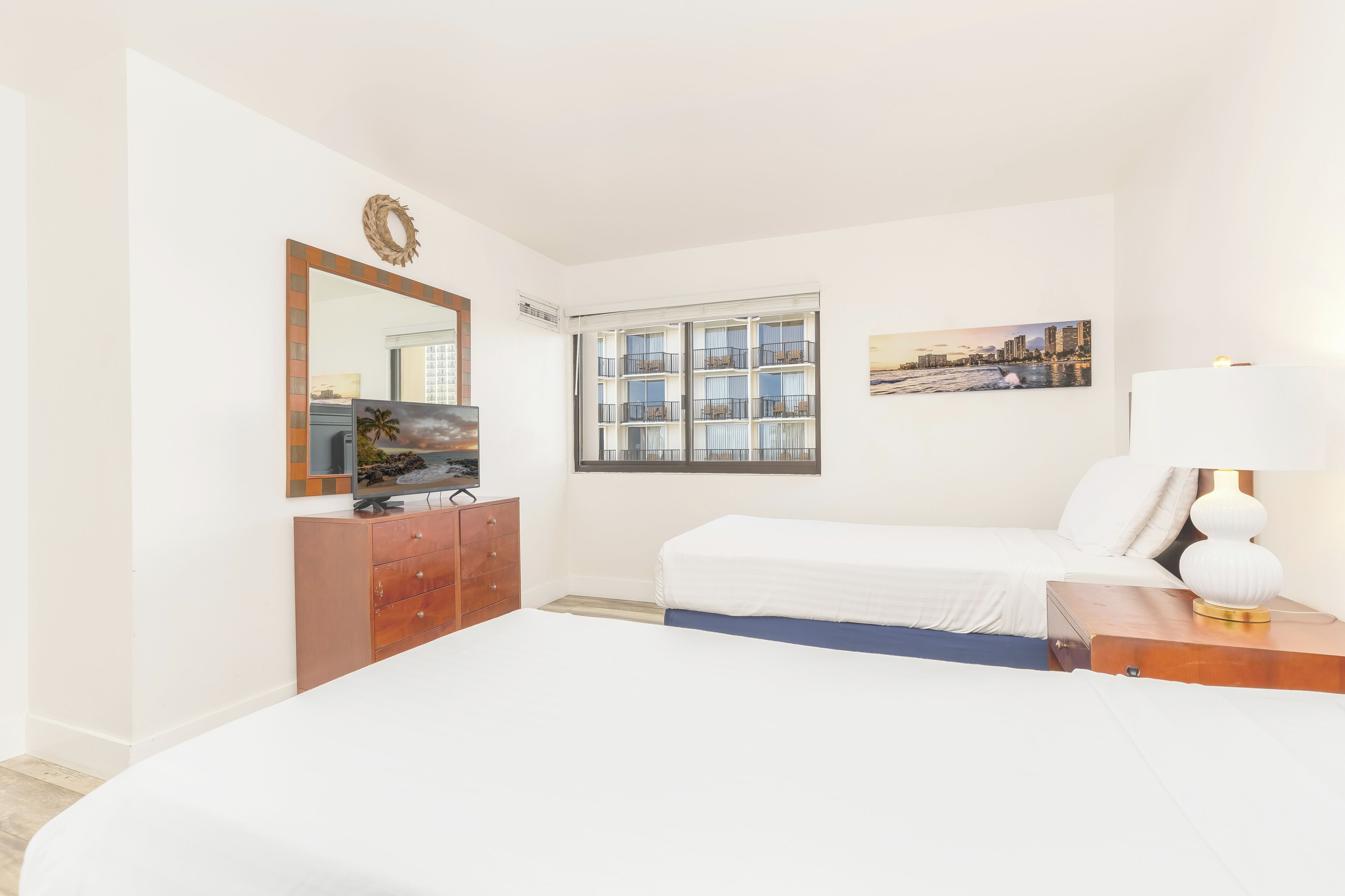Une chambre d’hôtel avec un grand lit, une commode, un miroir, une fenêtre donnant sur des immeubles et un tableau accroché au mur.