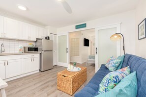 A modern kitchen with white cabinets, a refrigerator, and a microwave. There is a wicker coffee table with a tray and candles. A blue sofa with colorful cushions is in the living area. - Remodeled Condo by the Beach at the Cabana Waikiki W/full Kitchen & Free Wifi (Honolulu)