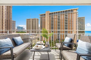 A balcony with wicker furniture, a table with a bottle and glasses, and a view of high-rise buildings. - Ocean & Fireworks Views Condo at the Ilikai Hotel W/full Kitchen & Free Wifi (Honolulu)