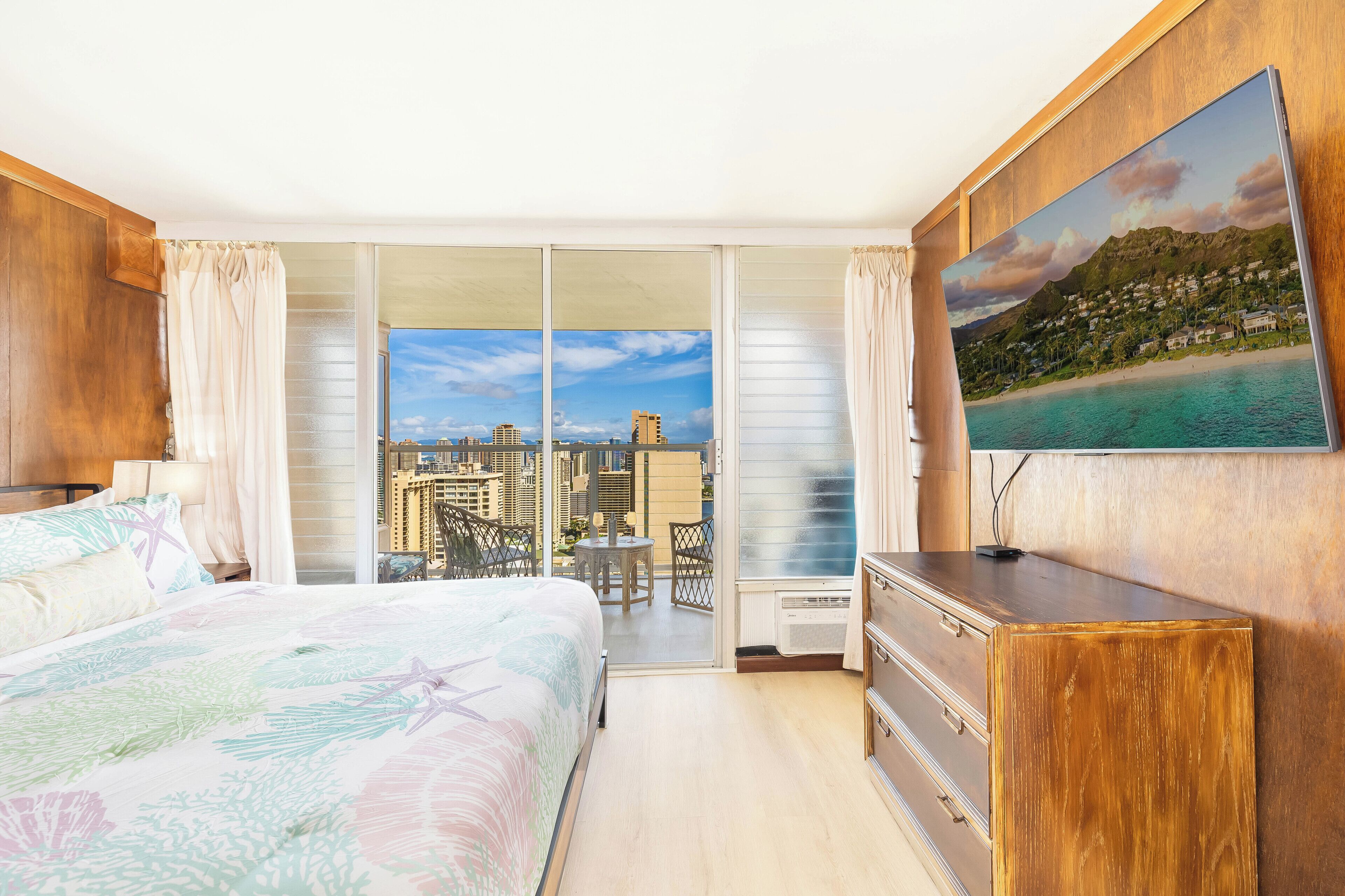 A hotel room with a bed, a desk, a chair, a TV, and a balcony with a city view.