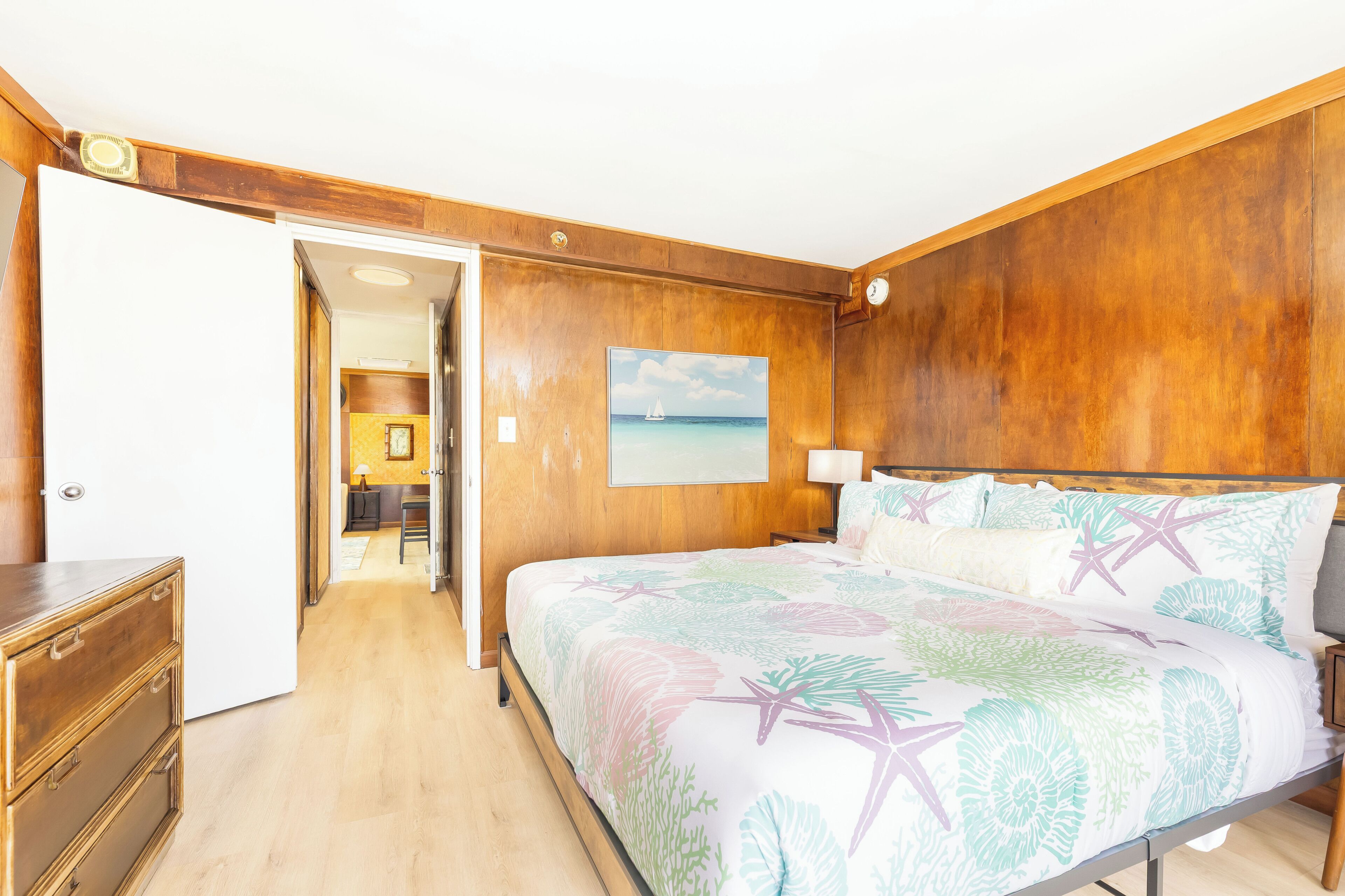 Une chambre à coucher avec une tête de lit en bois, un lit recouvert d’une literie à motifs floraux, une table de chevet, une lampe et un tableau représentant un voilier accroché au mur.
