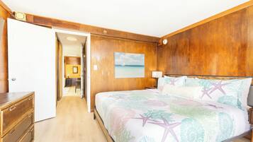 Une chambre à coucher avec une tête de lit en bois, un lit recouvert d’une literie à motifs floraux, une table de chevet, une lampe et un tableau représentant un voilier accroché au mur.
