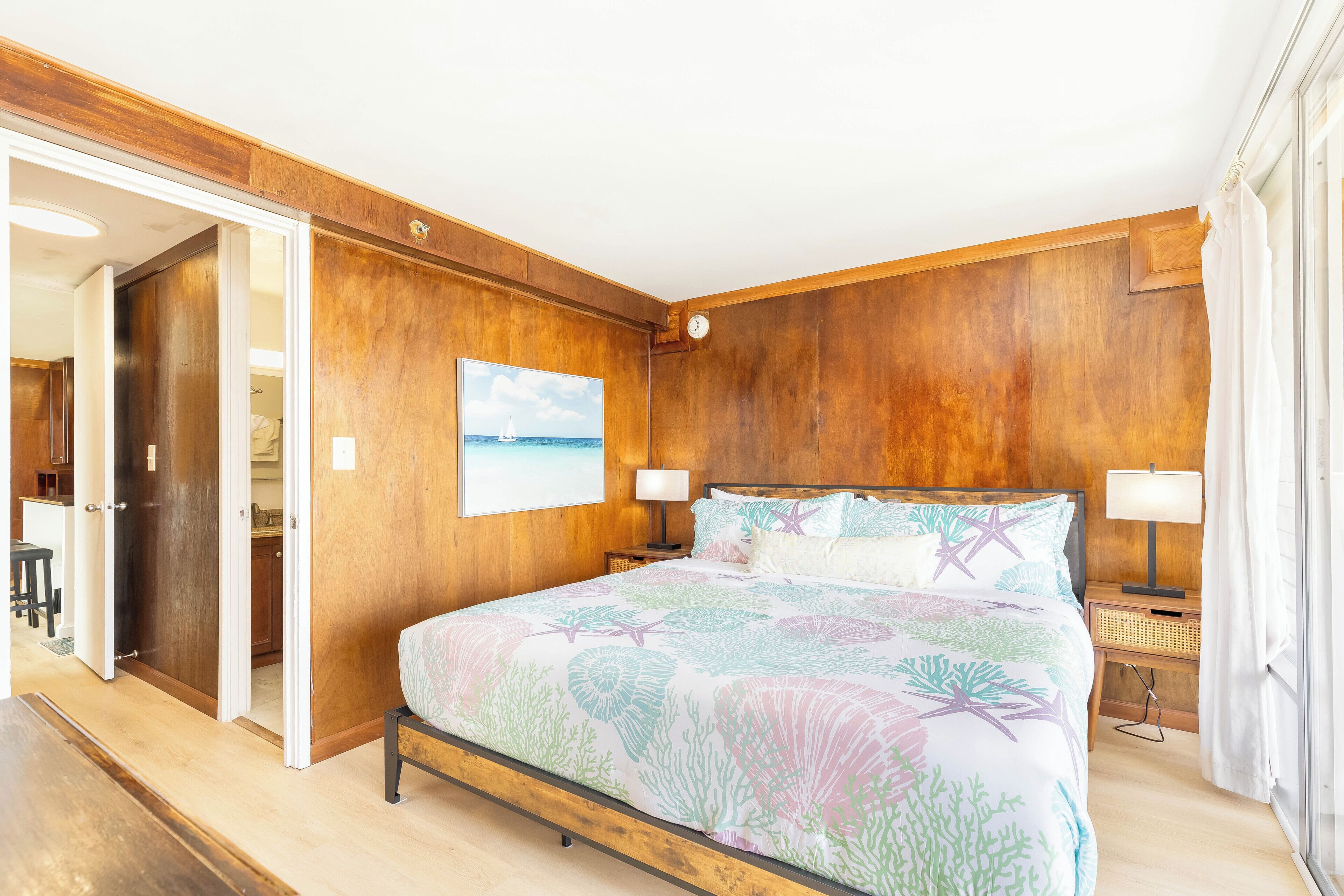A bedroom with a large bed, wooden walls, a painting, and a view of the kitchen.