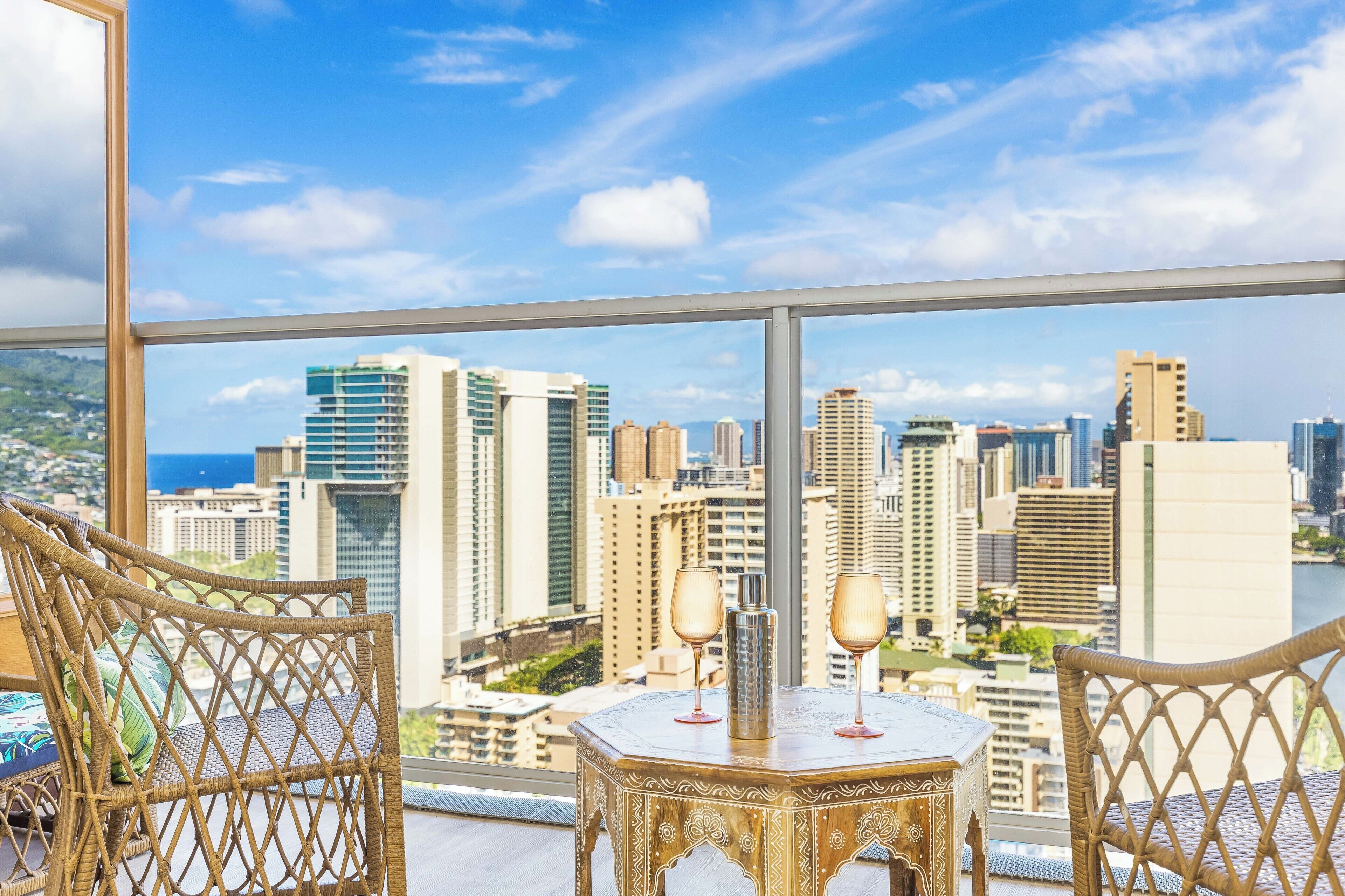 A balcony with wicker chairs, a table with two wine glasses, and a cityscape view.