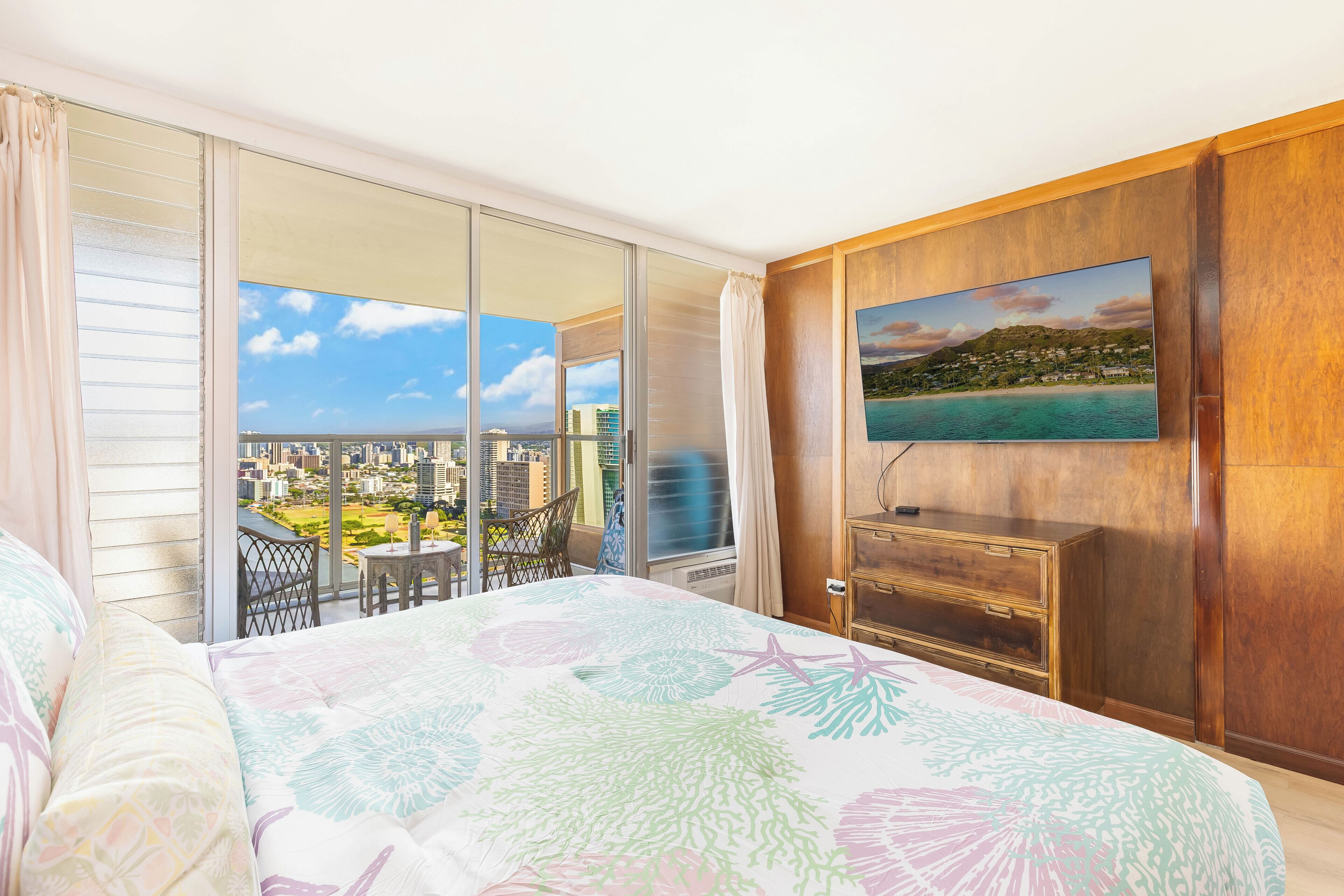 A hotel room with a large bed, a TV mounted on the wall, a balcony with a city view, and a wooden dresser.