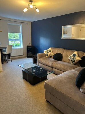 Ein Wohnzimmer mit einer Couch, einem Glastisch, einem Sessel, einer Heizung, einem Fenster und einer Deckenleuchte.