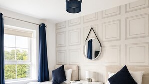 Ein Hotelzimmer mit zwei Betten, einem fensterbelaubten Fenster, einem Nachttisch mit Lampe und einem an der Wand befestigten Spiegel.