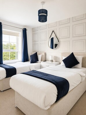 A hotel room with two beds, a window with curtains, a bedside table with a lamp, and a wall-mounted mirror. - Modern 3-Bedroom Home in Wakefield – Sleeps 5 (Wakefield)