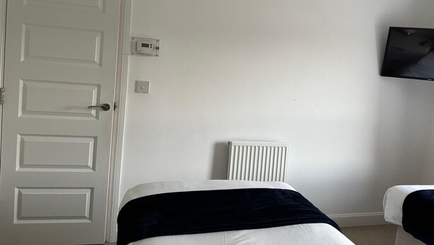 Ein Zimmer mit einer weißen Tür, einem an der Wand montierten Fernseher, einem Heizkörper und einem Bett mit dunkler Decke.
