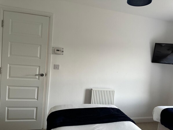 A room with a white door, a wall-mounted TV, a radiator, and a bed with a dark blanket. - Modern 3-Bedroom Home in Wakefield – Sleeps 5 (Wakefield)