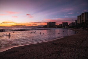 A beach with people swimming and walking along the shore, buildings in the background, and a sunset sky. - Marine Surf Waikiki Condo Short Walk to the Beach W/full Kitchen & Parking (Honolulu)