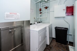 Une buanderie avec une armoire métallique portant l’inscription « DÉCHETS », un lave-linge blanc, une poubelle noire et un détecteur d’incendie au mur.