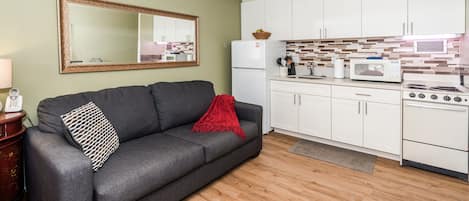 A compact living space with a grey sofa, a red throw blanket, a wooden floor, a kitchenette with white cabinets, a microwave, and a stove.