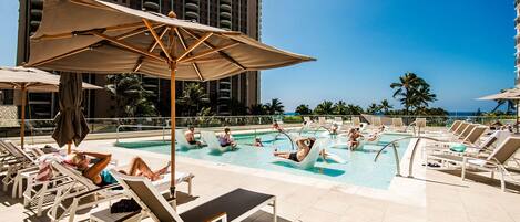 A poolside area with lounge chairs and umbrellas, a swimming pool, and a tall building in the background.