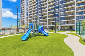 A playground with slides and climbing structures, surrounded by a well-maintained lawn and a large building with multiple balconies. - Diamond Head View Rare Condo at the Waikiki Banyan by the Beach With Parking (Honolulu)
