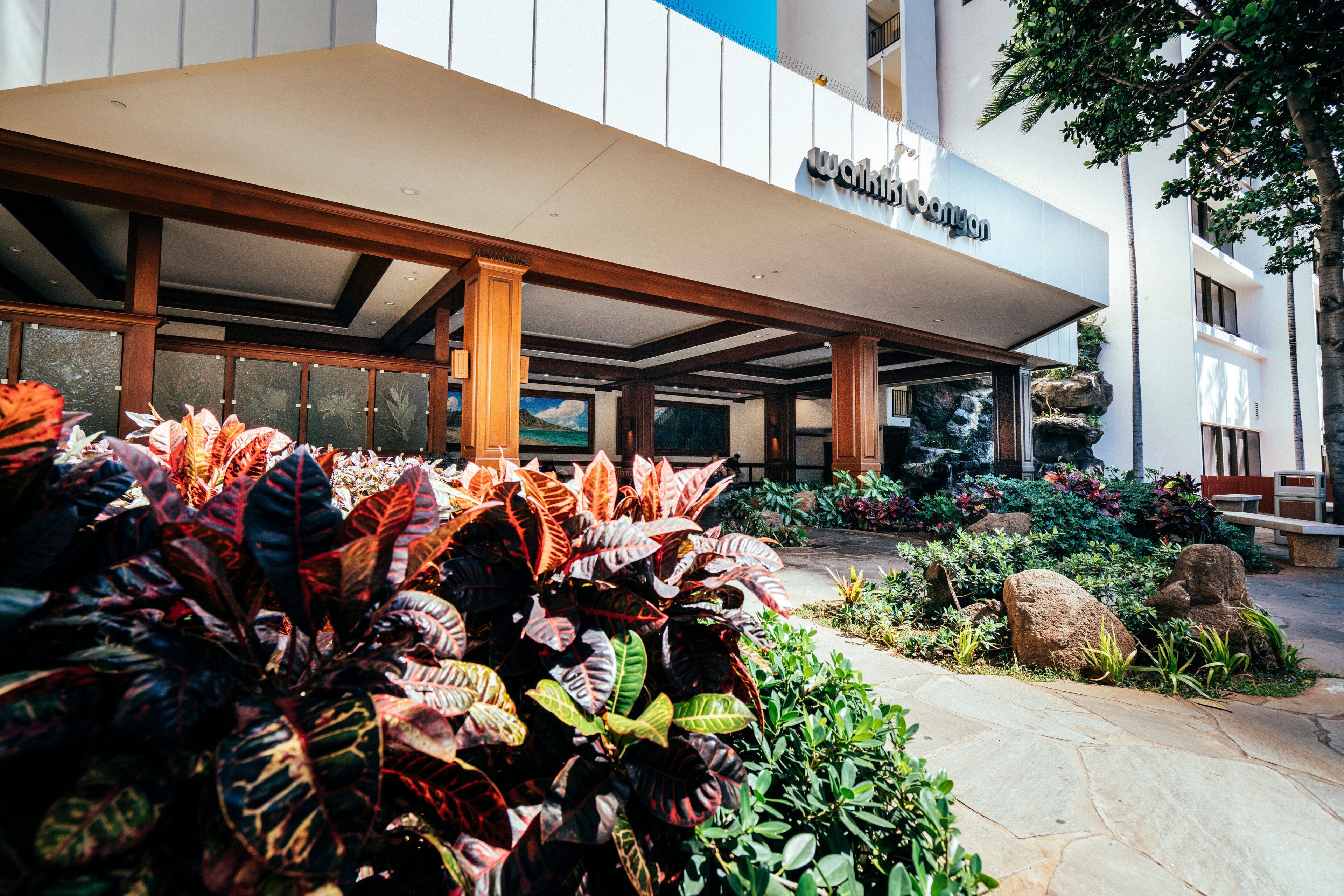 A hotel entrance with a modern facade, a landscaped garden with tropical plants, and a clear view of the building's entrance.