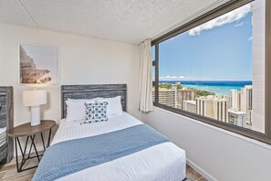A hotel room with a bed, a nightstand, a lamp, a framed picture, and a large window with a city and ocean view. - Ocean View Fully Renovated Condo at Waikiki Banyan W/amazing Amenities (Honolulu)