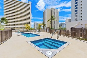 A rooftop pool area with two pools, surrounded by tall buildings, palm trees, and a clear sky. - Ocean View Fully Renovated Condo at Waikiki Banyan W/amazing Amenities (Honolulu)