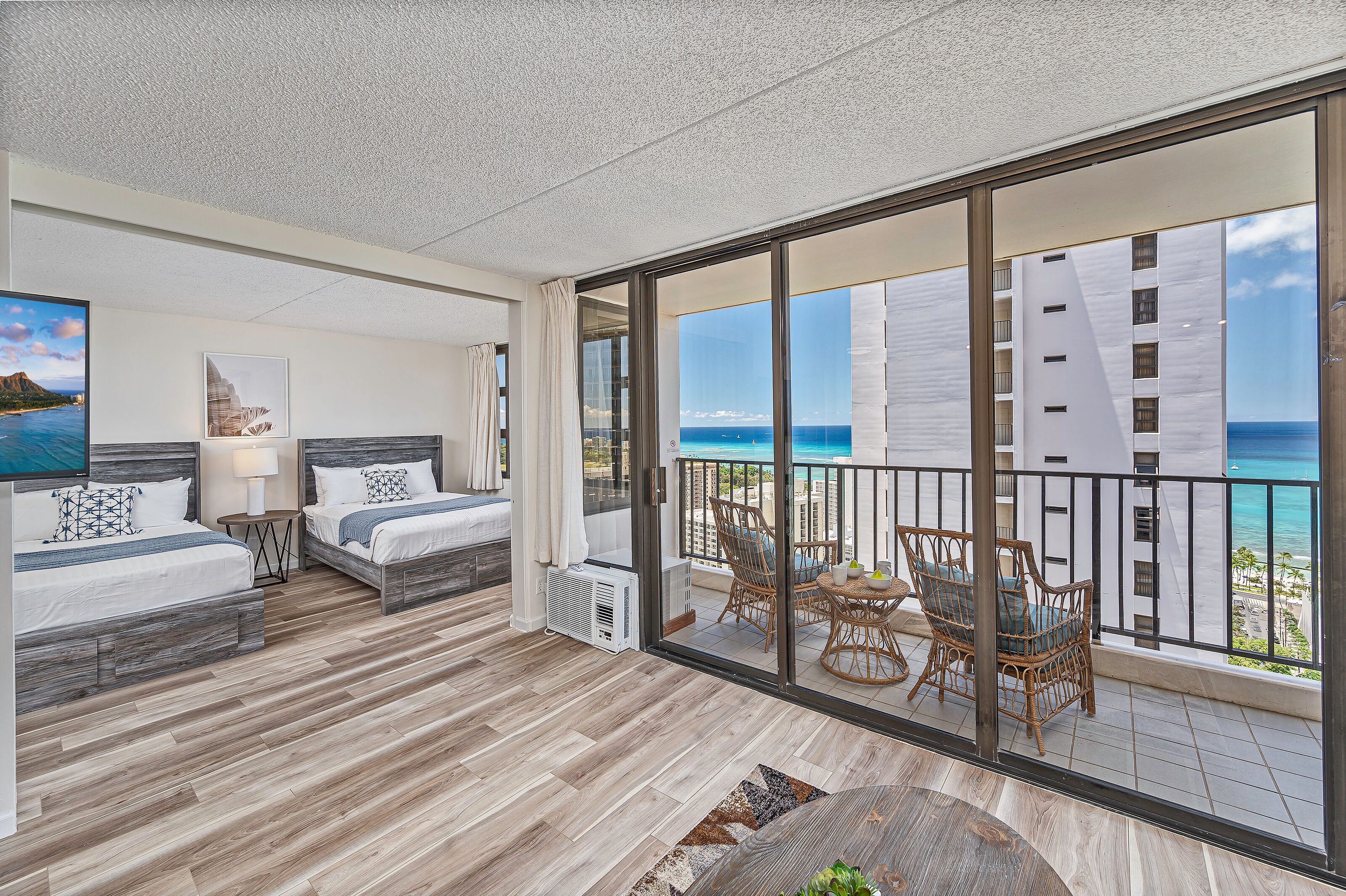 A balcony with a view of the ocean and a building. There are two beds with white bedding and a small table with a lamp.