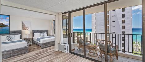 A balcony with a view of the ocean and a building. There are two beds with white bedding and a small table with a lamp.