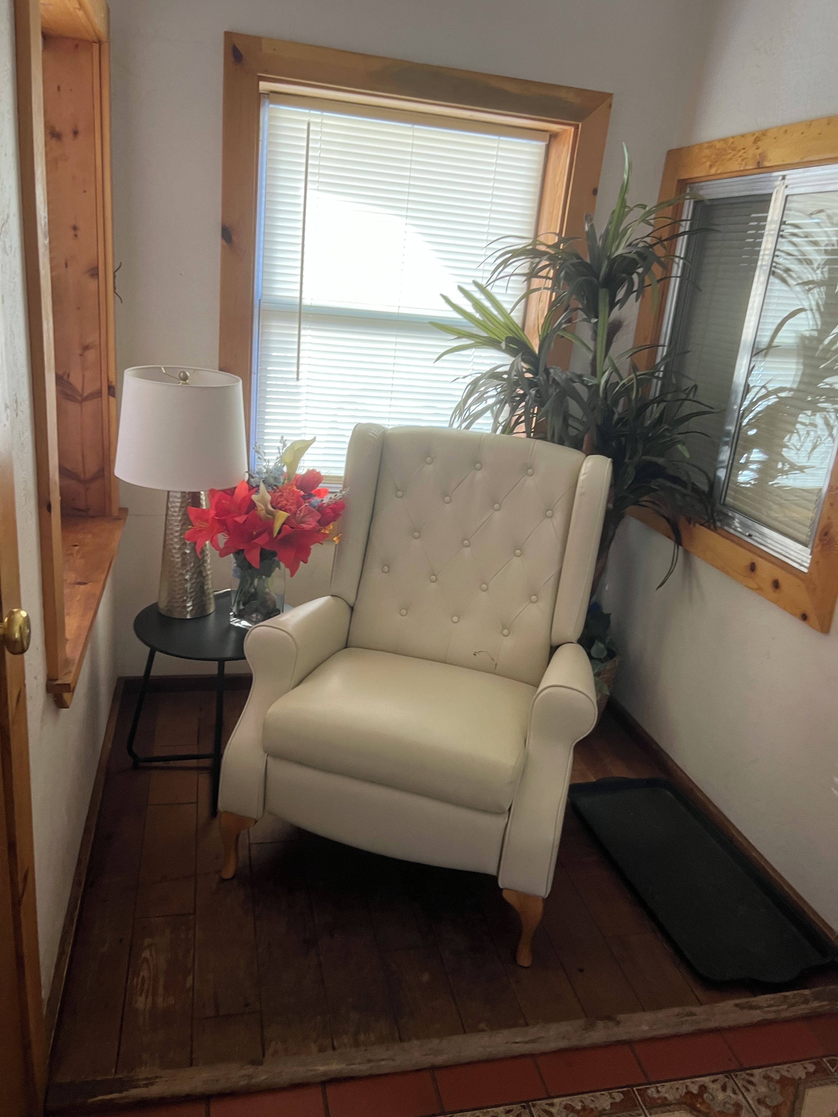 Una habitación de esquina con un sillón color beige, una mesita con una lámpara, una planta en maceta y una ventana con persianas.