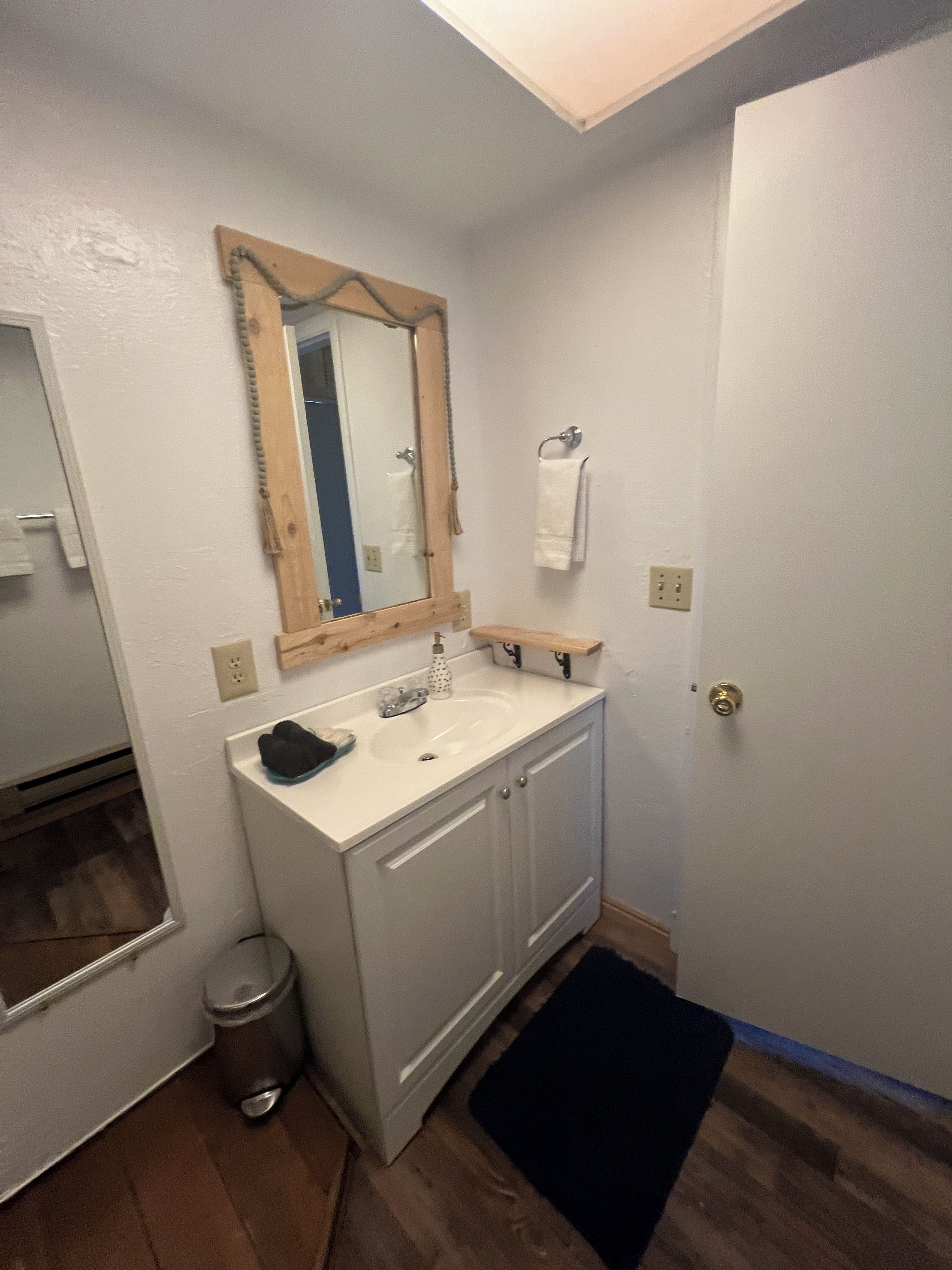 Un baño con un lavamanos blanco, un espejo con marco de madera, una toalla colgada en la pared y una puerta.