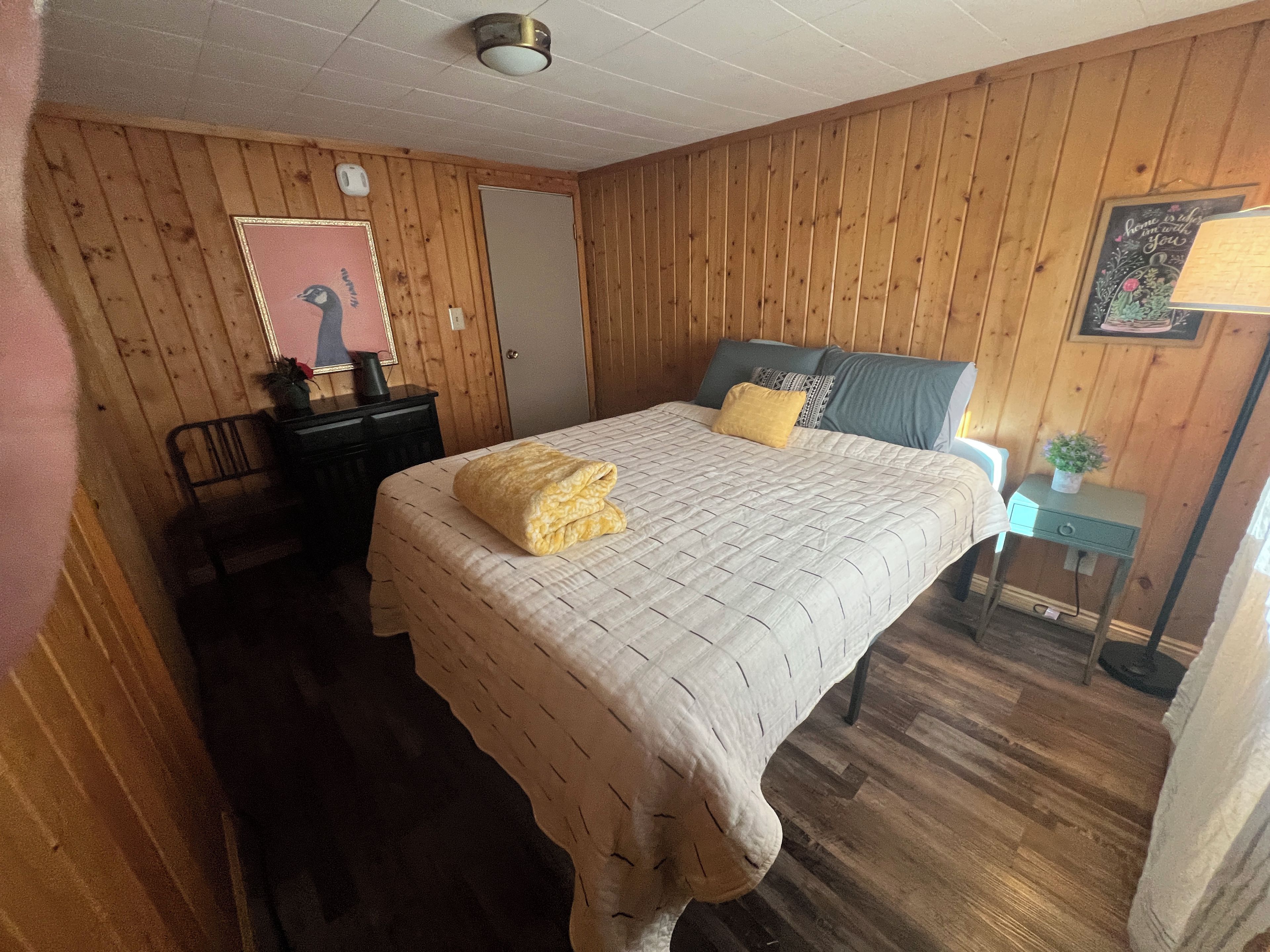Una stanza con un letto, una scrivania, una sedia, un tavolino con una pianta, e un muro decorato da un quadro e una targa.
