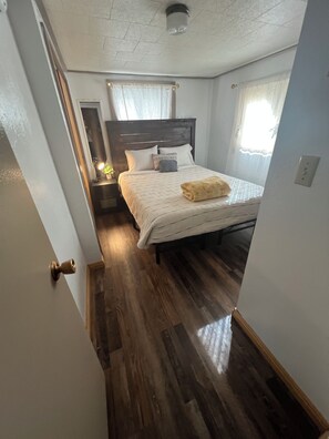 A bedroom with a wooden floor, a bed with a white bedspread and pillows, a nightstand with a lamp, and a window with sheer curtains.