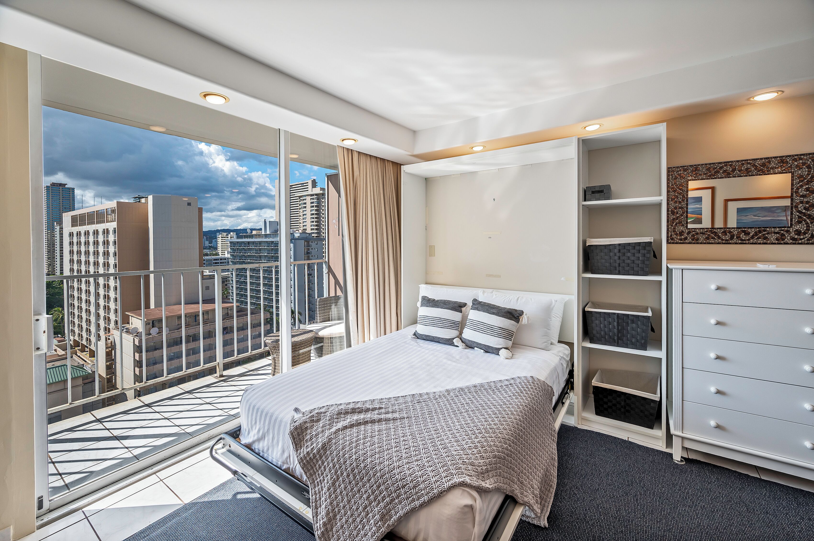 A modern hotel room with a balcony, a bed with a patterned bedspread, a dresser, and a large mirror.