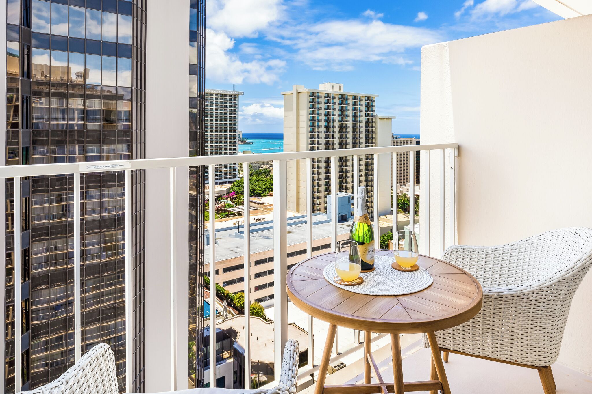 A balcony with a round table, wicker chairs, a bottle of champagne, and a glass of orange juice.