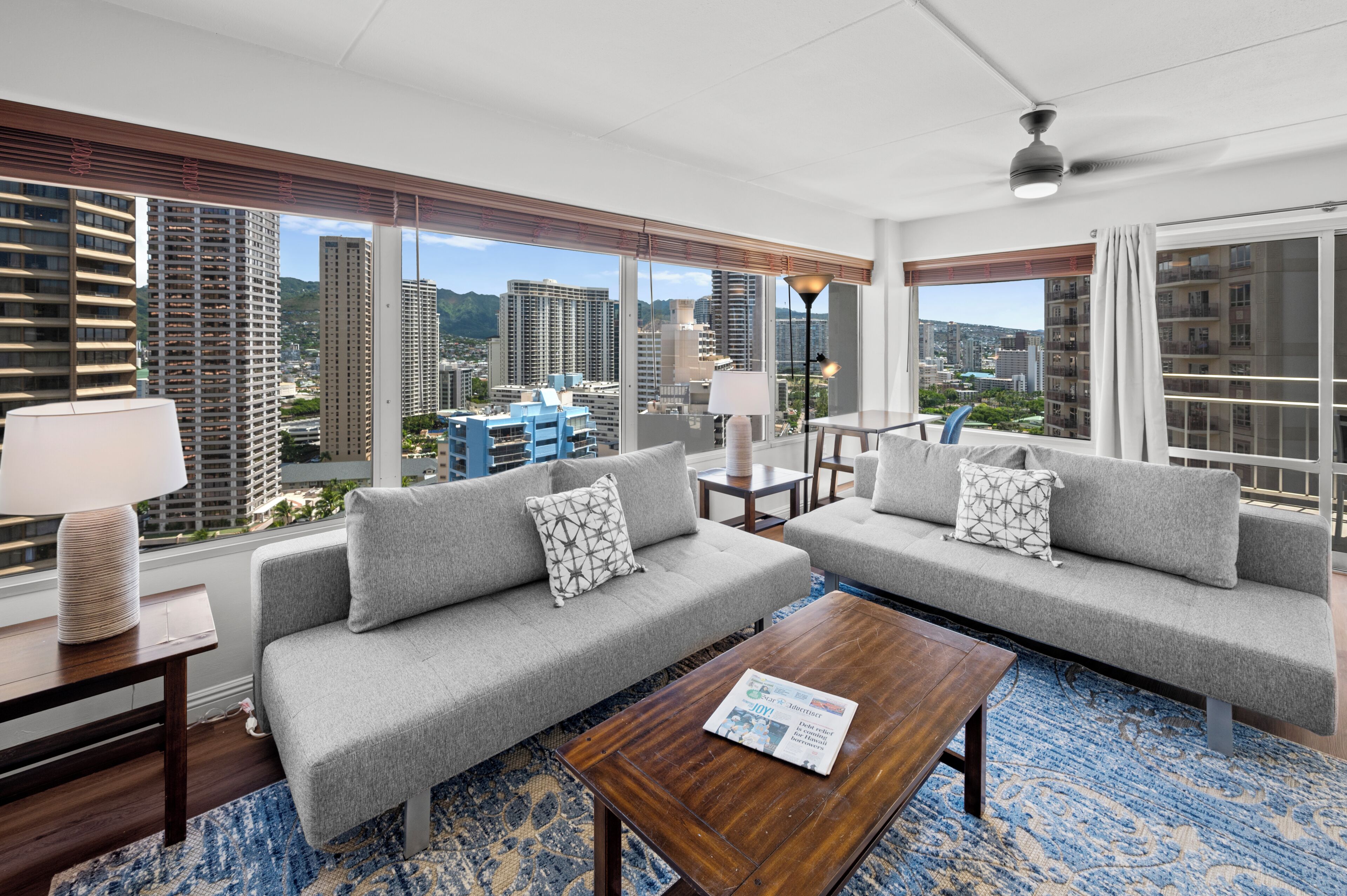 A modern hotel room with two grey sofas, a wooden coffee table, and a large window offering a city view.