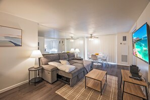 A modern living room with a sectional sofa, a small round table, a coffee table, a TV, and a dining area with a white table and chairs. - Ocean-marina Yacht Views Condo Steps to the Beach W/full Kitchen & Free Wifi (Honolulu)