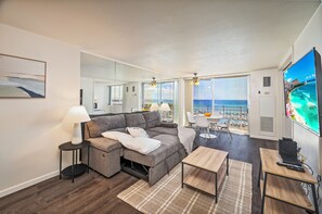 A modern living room with a gray sectional sofa, a small coffee table, a TV, and a view of the ocean through large windows. - Ocean-marina Yacht Views Condo Steps to the Beach W/full Kitchen & Free Wifi (Honolulu)