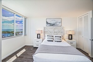 Una camera da letto con un'ampia finestra vista mare, un letto con copriletto grigio e bianco, due comodini con lampade e una testiera in legno.