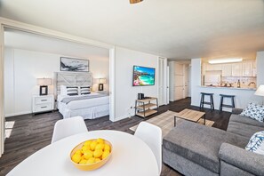 Un soggiorno moderno e luminoso con un divano grigio, un tavolo da pranzo bianco con una ciotola di frutta, un letto con biancheria da letto bianca e una TV a parete.
