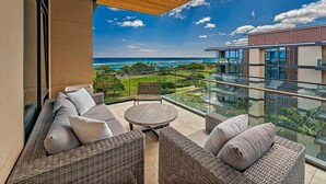 Ein Balkon mit Korbmöbeln, einem runden Tisch und einem Glasgeländer mit Blick auf eine Küstenlandschaft.