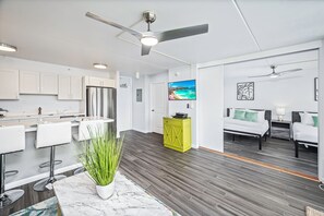 Eine moderne Küche mit weißen Schränken, einem Edelstahlkühlschrank und einer Bar mit weißen Hocker. Im angrenzenden Raum befindet sich ein grüner Schrank und ein Bett mit weißer Bettwäsche.