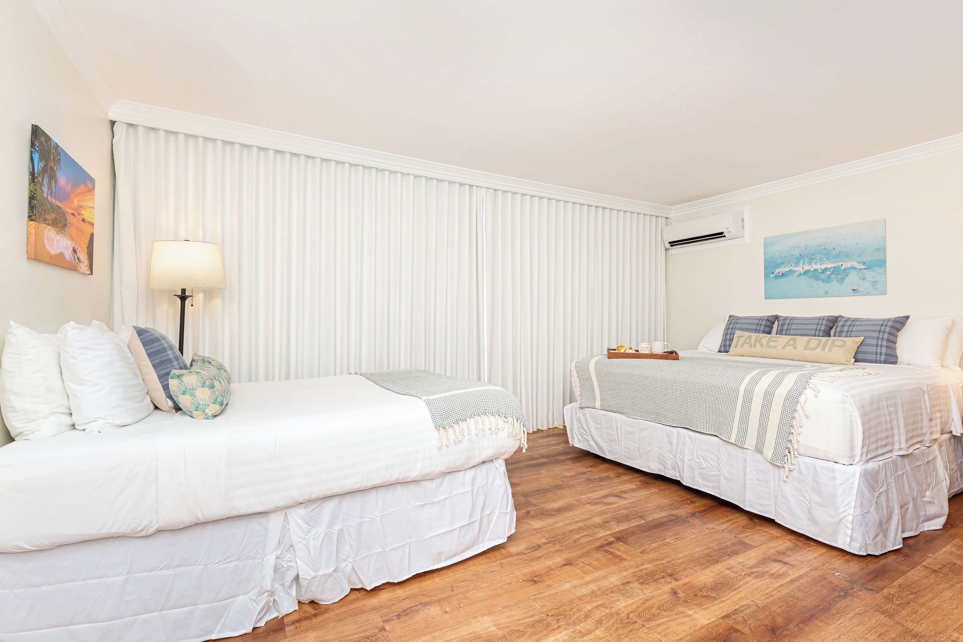 Une chambre d’hôtel avec deux lits, une grande fenêtre avec stores, un climatiseur fixé au mur et un tableau.